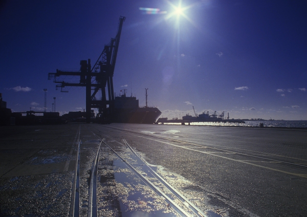 A sunlit dockyard with large dockside cranes in the background