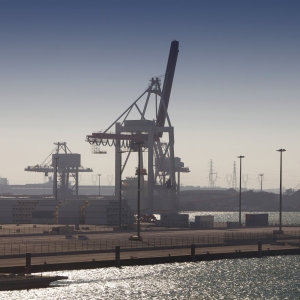 Cranes and containers at a modern dock terminal, with older heavy industry installations in the background