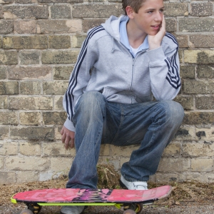 A young teenage boy squatting against an urban wall with bricks and graffiti and skateboard