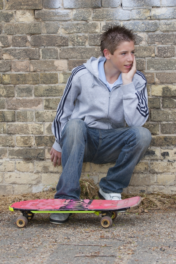 A young teenage boy squatting against an urban wall with bricks and graffiti and skateboard